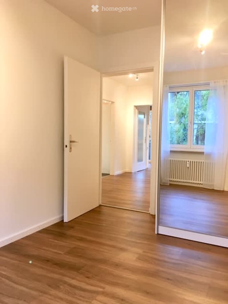 Empty room with light wooden flooring, white walls, and a partially open door leading to another room. Natural light from a window on the right side.