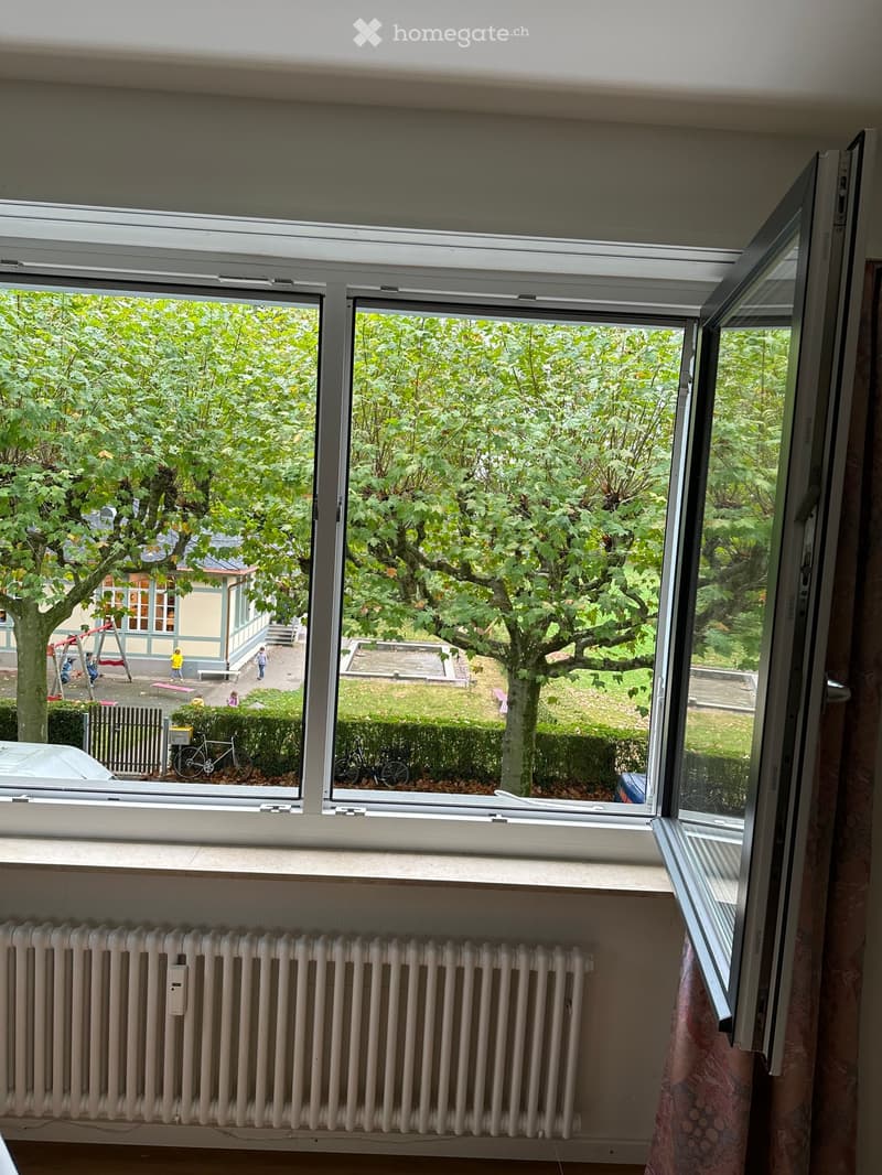 An unfurnished room with large windows offering a view of a park-like area with trees, a playground, and a house in the background. The room features a radiator and a partially visible curtain.