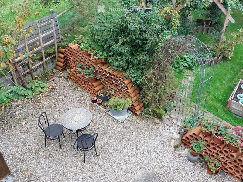 pebble area with table and chairs, green garden, rows of terracotta pots, arch trellis, wooden fence, grassy area, tree