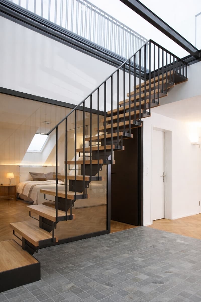modern staircase with wooden steps and black metal railings, glass wall, wooden floor, bedroom with bed and lamp