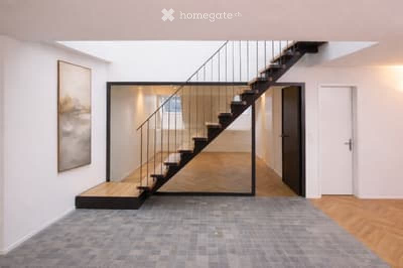 Modern hallway with a sleek staircase featuring a glass railing, grey carpeted floor, and wooden paneling on the walls.