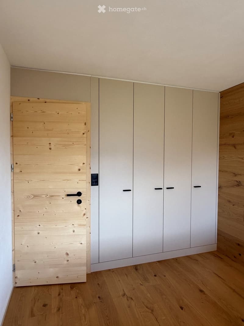 Empty room with light wooden flooring, white walls, and built-in white cabinets with black handles. A wooden door is visible on the left side.