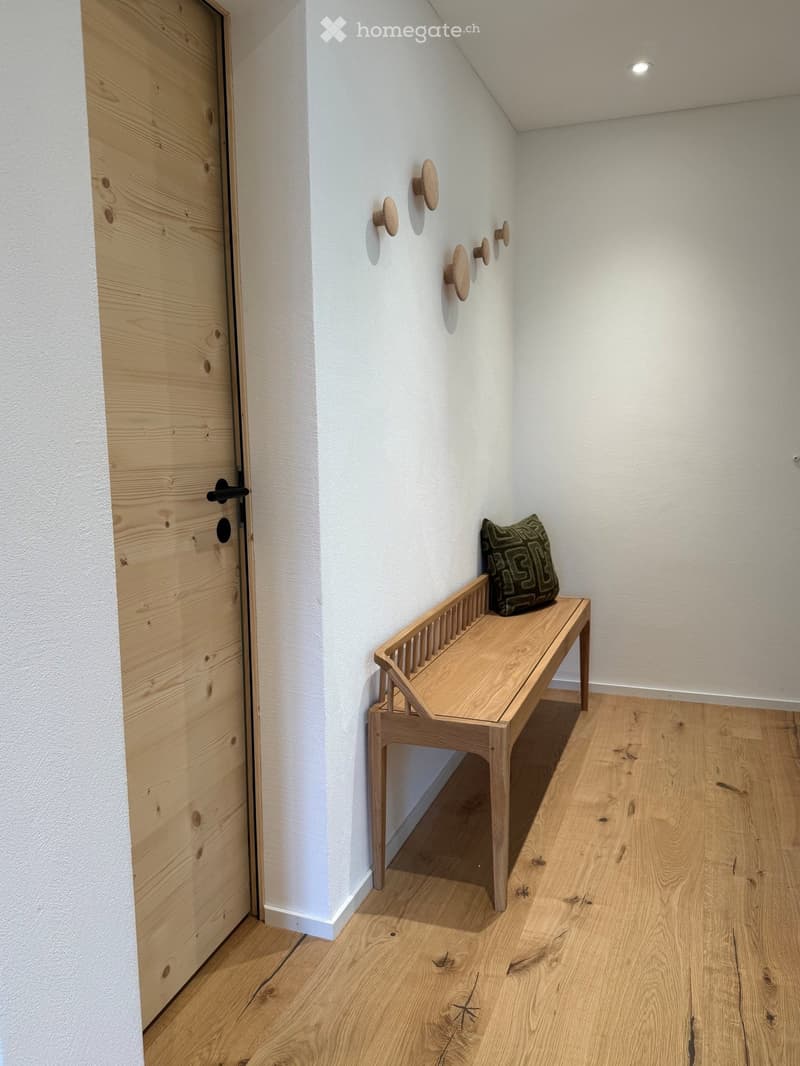 A narrow hallway with light wooden flooring, white walls, a wooden door on the left, and a small wooden bench with a decorative pillow on the right.