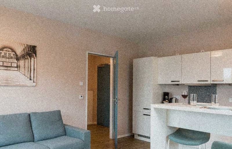 A modern, compact living space with a sofa, coffee table, and a small kitchen area featuring cabinets, a countertop, and various items including a wine glass and coffee maker.