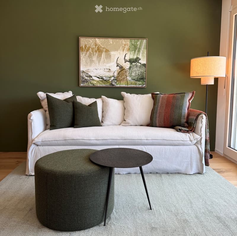 Living room with a white sofa, green and white pillows, a round green stool, a black coffee table, a floor lamp, and a framed painting on the wall.