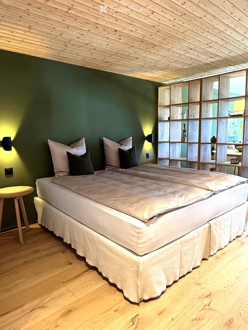 Cozy bedroom with a large double bed, white bedding, dark green pillows, wooden floor, and a small round wooden stool next to the bed.