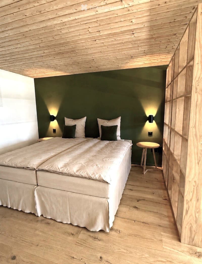 Cozy bedroom with wooden ceiling and floor, double bed with white bedding and black pillows, small wooden stool, and wall-mounted lights.