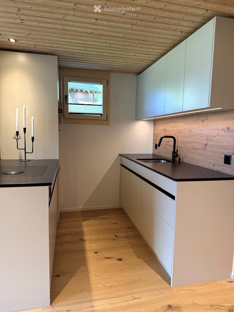 Modern kitchen with light wooden cabinets, dark countertops, and a sleek sink with a modern faucet. A window provides natural light, and a candle holder with candles sits on the counter.