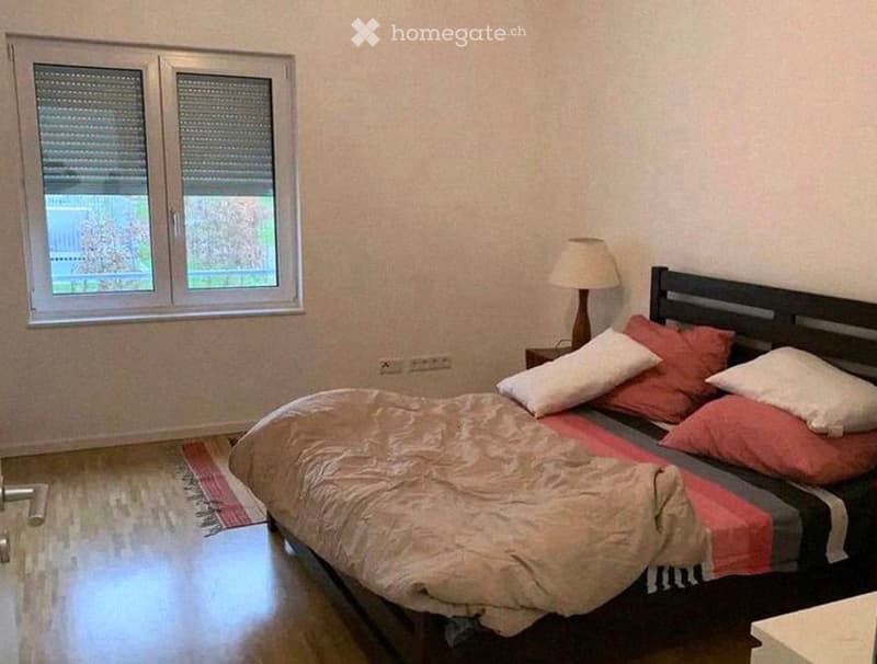 Cozy bedroom with a double bed, pillows, a nightstand, and a lamp. The room features wooden flooring and a window with blinds.