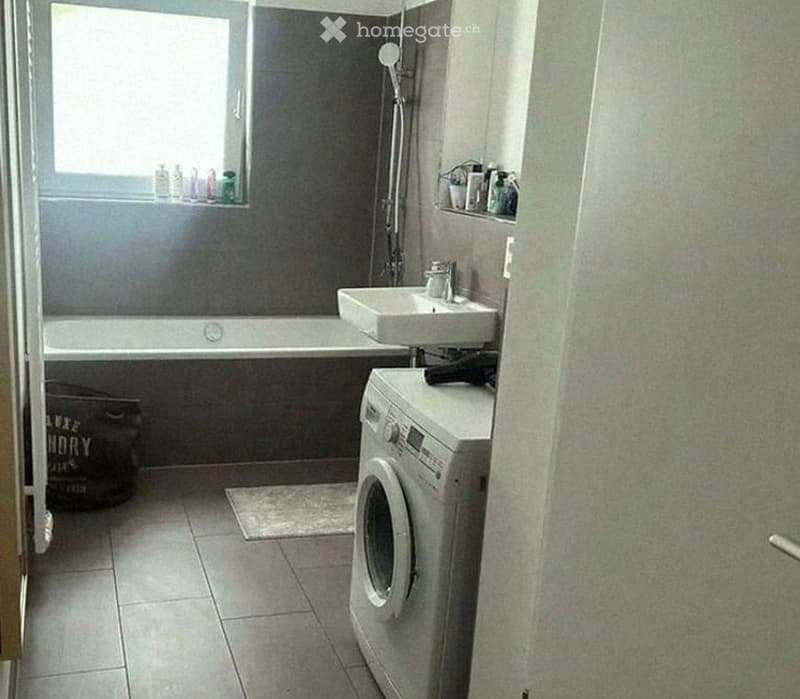 Bathroom with a bathtub, sink, washing machine, and shower area. The space is tiled and has a window with toiletries on the sill.