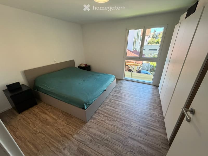 Bedroom with a single bed, nightstand, and a small table near the window. The room has wooden flooring and white walls.