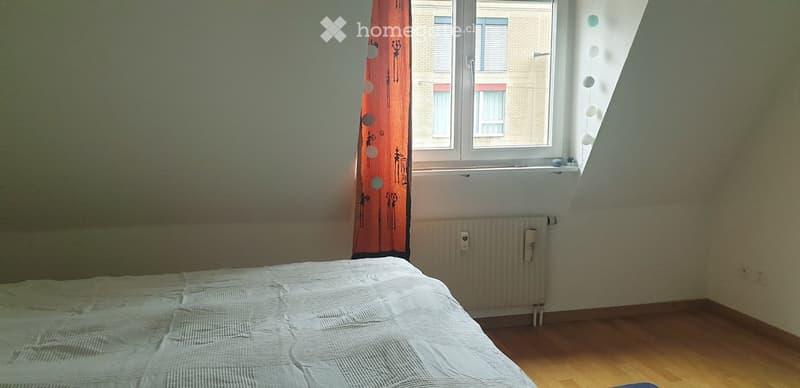Bedroom with a single bed, wooden floor, window with orange curtain, radiator, and visible cityscape through the window.