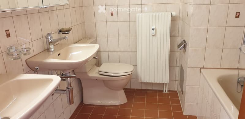 Bathroom with beige tiles, a sink with a chrome faucet, a toilet, a bathtub, a radiator, and a mirror above the sink. There are two glass cups on the wall next to the sink.