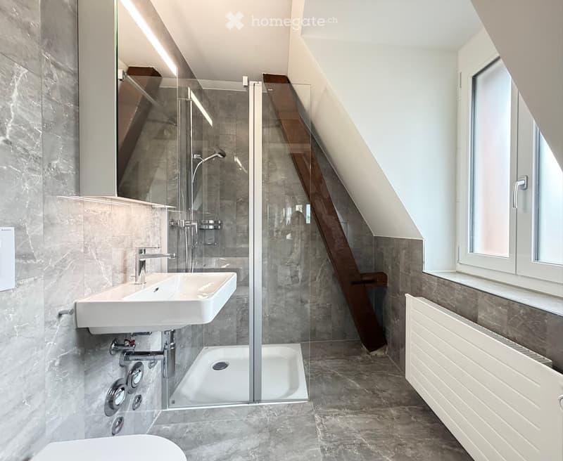 Modern bathroom with a white sink, chrome faucet, glass-enclosed shower, and radiator. The walls are tiled in grey marble, and the floor is made of dark stone tiles.