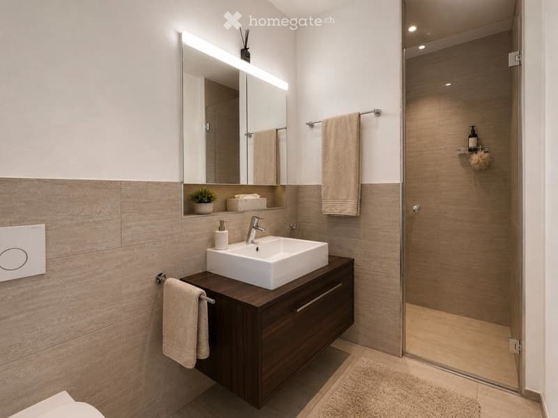 Modern bathroom with a white sink, dark wooden vanity, large mirror with LED lighting, and a glass-enclosed shower. Beige tiles and towels are visible.