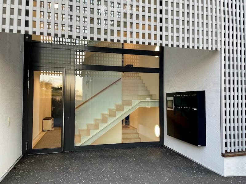 Modern building entrance with glass doors, visible staircase inside, raised ground floor, and mailboxes on the wall.