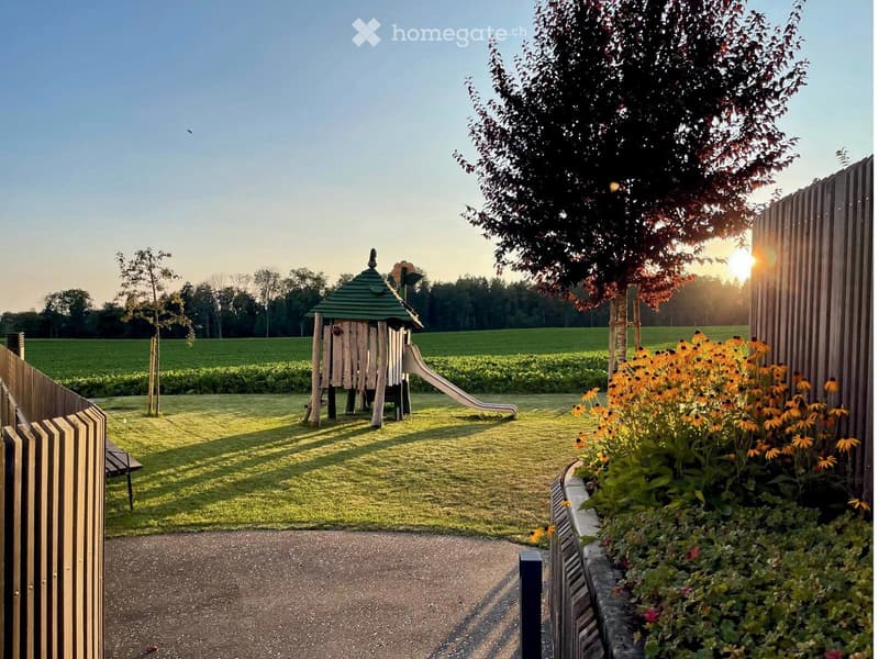 A well-maintained garden area with a wooden play structure, slide, and colorful flowers, surrounded by a wooden fence and open fields.