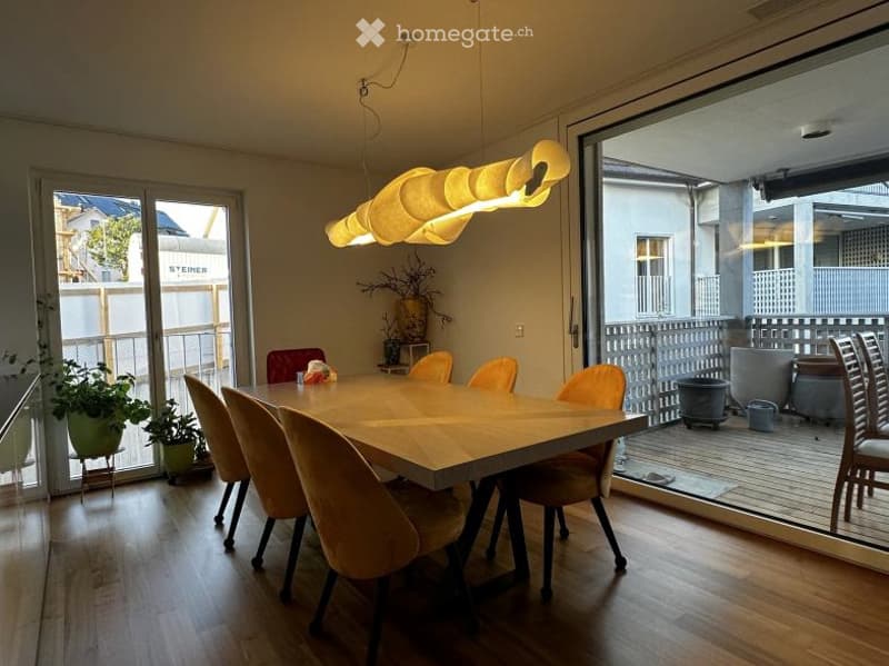 Bright dining area with a wooden dining table, six chairs, a modern hanging light fixture, and potted plants near large glass doors leading to a balcony.