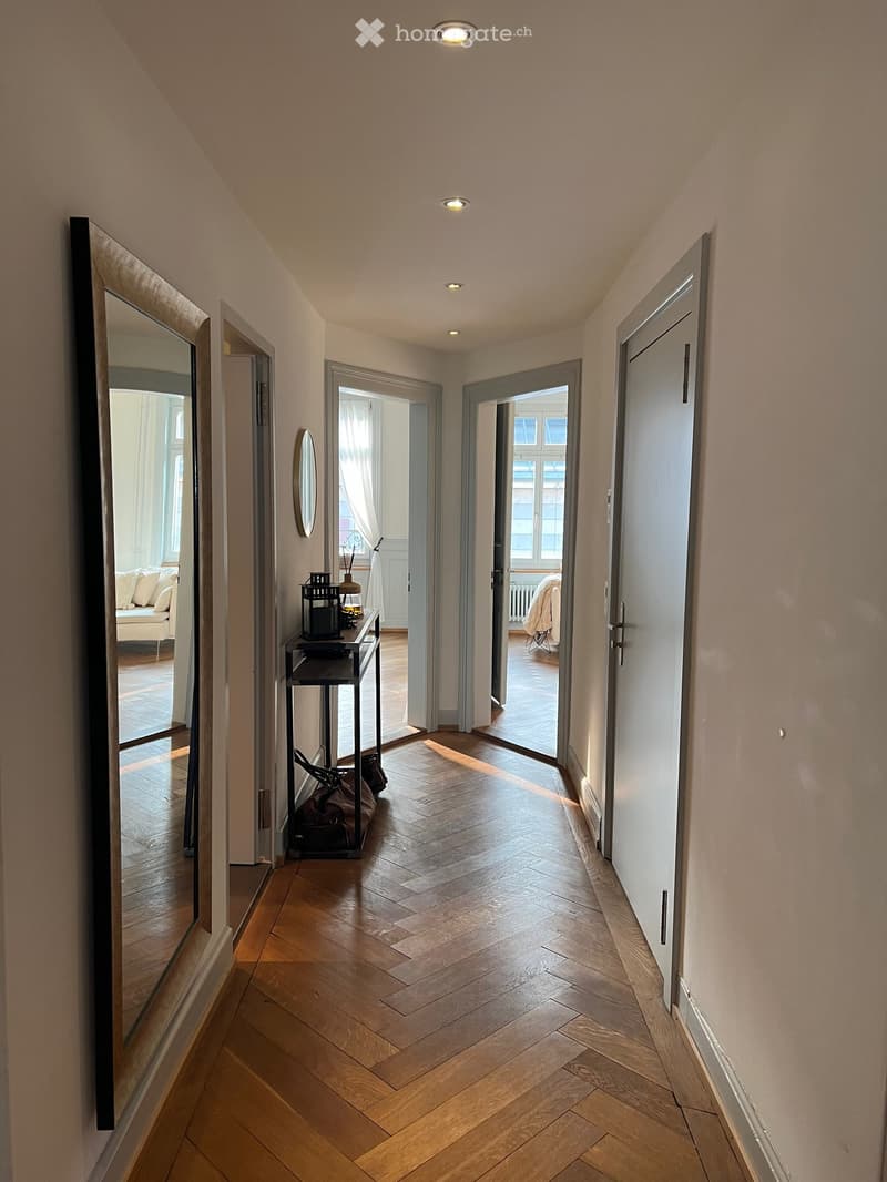A well-lit hallway with wooden herringbone flooring, several doors, and recessed ceiling lights. A small table with a lamp and decorative items is visible to the left.