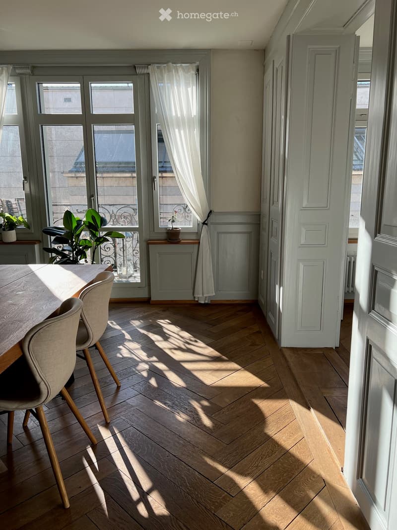 Bright dining area with wooden herringbone flooring, large windows with white curtains, and a dining table with chairs.