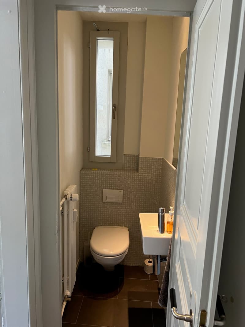 Modern bathroom with a toilet, sink, and small window. Features tiled walls, a towel, and a soap dispenser on the sink.