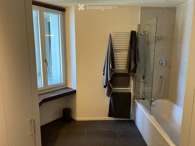 Modern bathroom with dark tiled floor, white walls, bathtub with glass shower enclosure, towel rack with towels, window with wooden sill, and a trash can.