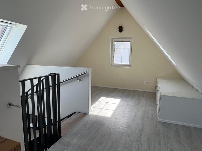 Bright, open hallway with wooden flooring, white walls, and a modern metal railing leading to a staircase. Features a skylight window and a smaller window on the wall.