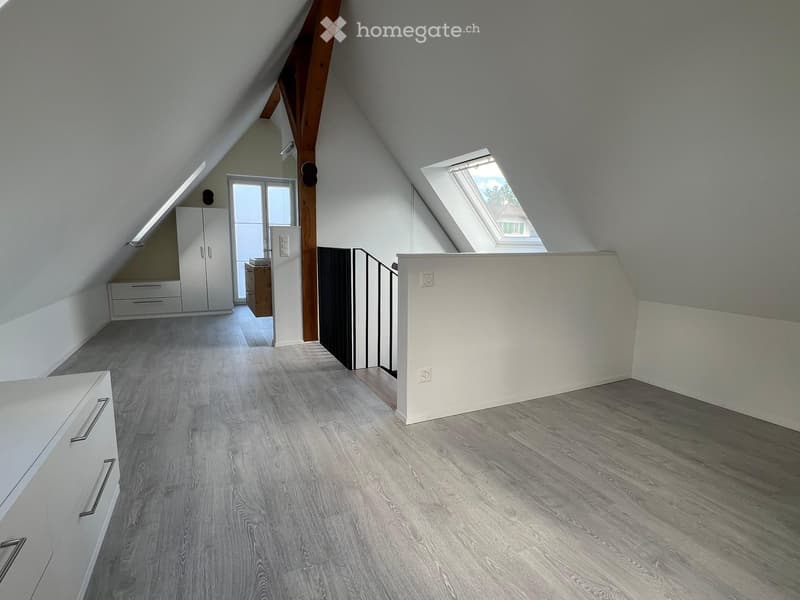 Bright, empty attic room with wooden flooring, white walls, and skylight windows. Features built-in white cabinetry and a small wooden desk area.