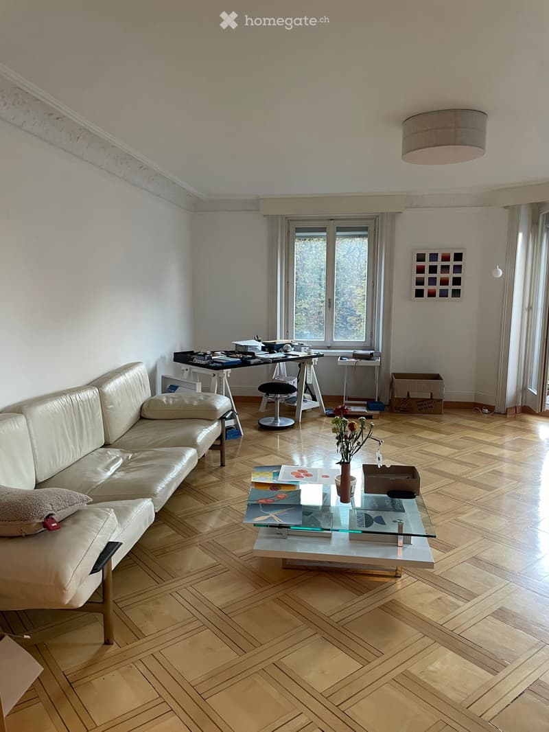 Living room with a cream-colored sectional sofa, glass coffee table, wooden floor, and a desk area with a chair and stool.