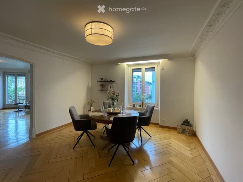 Dining room with a round wooden table, four chairs, a vase with flowers, and a shelf with decorative items. The room has wooden flooring and large windows with a view of the city.