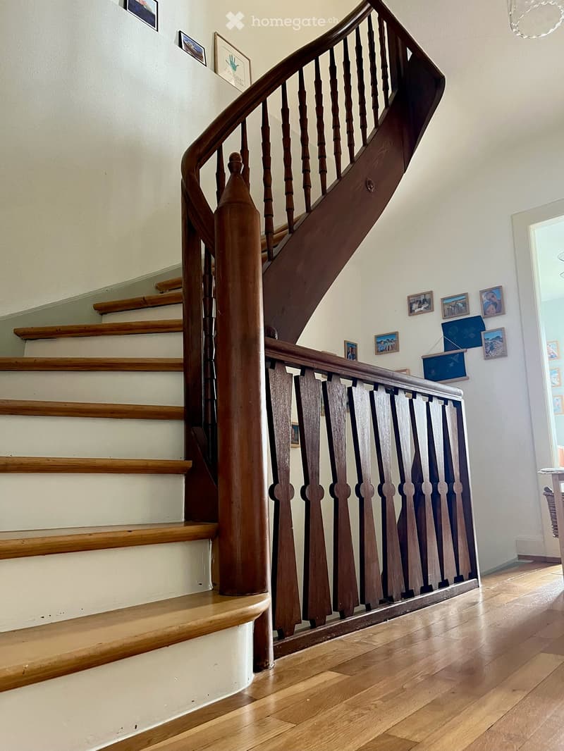 Wooden staircase with a polished handrail and balusters, leading to an upper level. The walls are painted white and decorated with several framed pictures. The floor is made of light wooden planks.