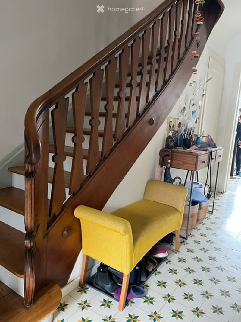 A hallway with a wooden staircase, a yellow bench, and a small table with items on it. The floor has a patterned tile design.
