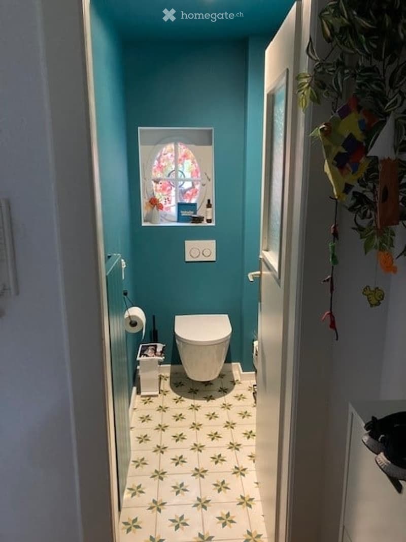A small bathroom with teal walls and a floral-patterned tile floor. Features a toilet, toilet paper holder, a small window with a decorative glass design, and a trash bin.