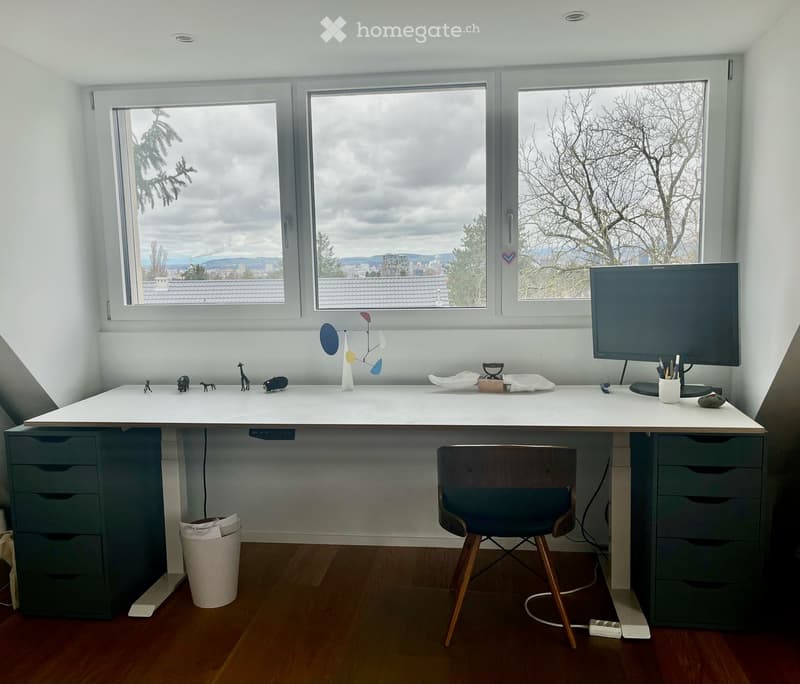A modern, well-lit room with a large desk, a comfortable chair, a screen, and various decorative items. The room features large windows with a scenic view of trees and rooftops.