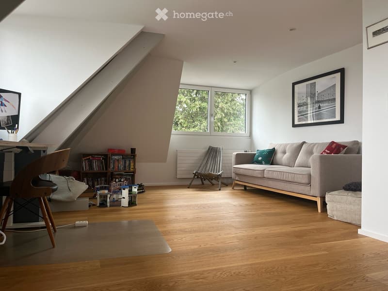 Bright living room with wooden flooring, large windows, a grey sofa with cushions, a wooden chair, a small table, a bookshelf, and a picture frame on the wall.