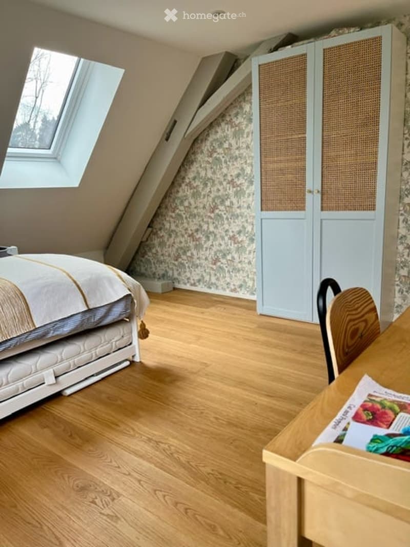 Cozy attic bedroom with wooden flooring, slanted ceilings, a skylight window, a bed with white bedding, a light blue wardrobe with wicker doors, and a wooden chair at a small table.