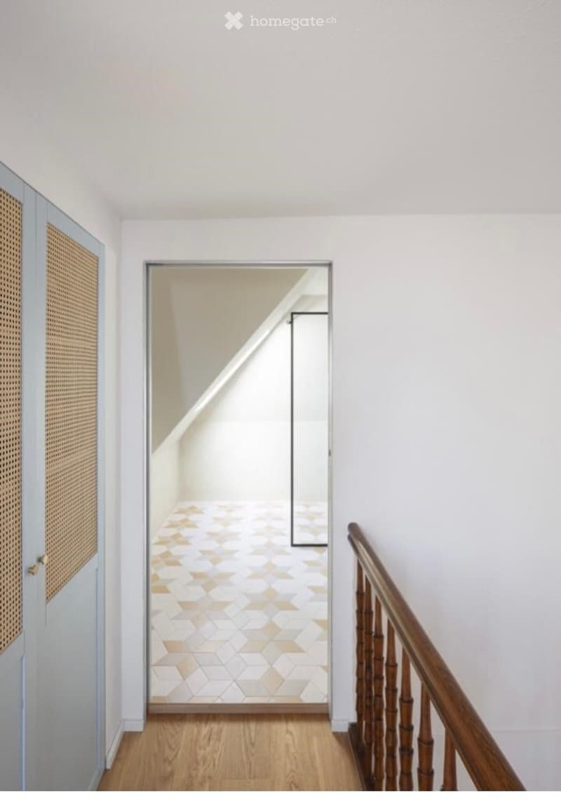 A modern hallway with white walls, wooden flooring, and a geometric patterned tile floor visible through an open doorway. Features a wooden railing and a woven door.