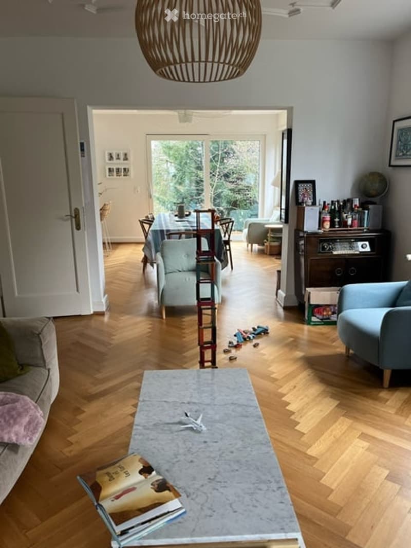 Open-plan living and dining area with wooden flooring, a grey sofa, a coffee table, armchairs, a dining table with chairs, a bookshelf, and a globe. The space is well-lit with natural light from large windows.