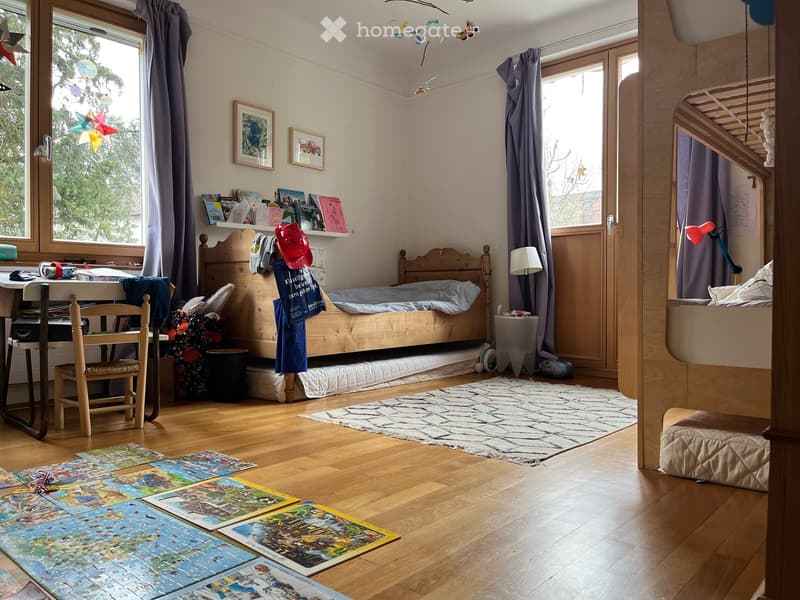 A well-lit bedroom with wooden floors, a bed with a wooden headboard, a small desk with a chair, a lamp, and several puzzles on the floor.