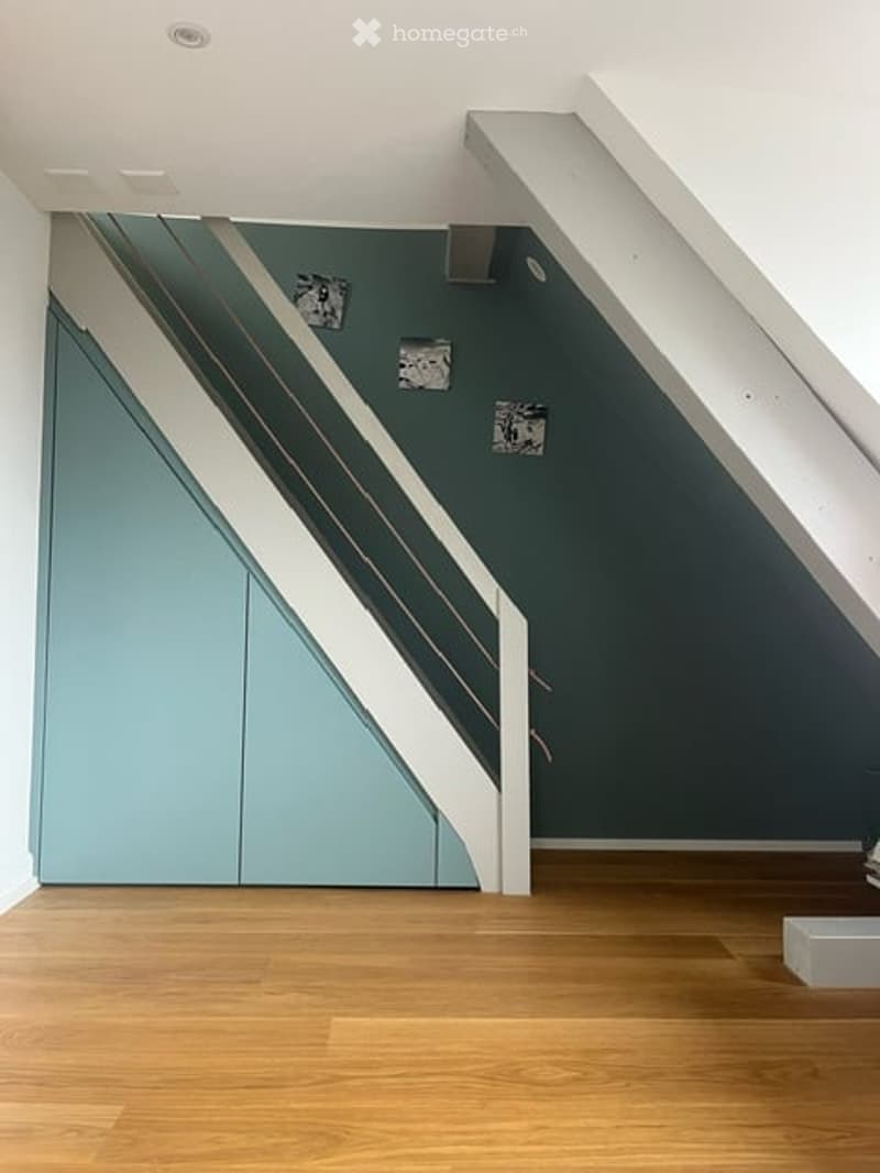 Empty room with wooden flooring, white ceiling, and a staircase with white railings and blue panels.