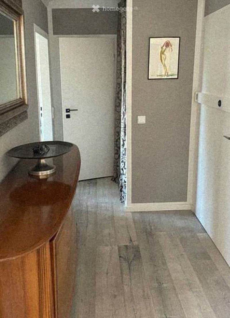 A narrow hallway with carpeted flooring, light-colored walls, and a wooden side table with a decorative item on top. A framed picture hangs on the wall, and a closed door is visible at the end of the hallway.