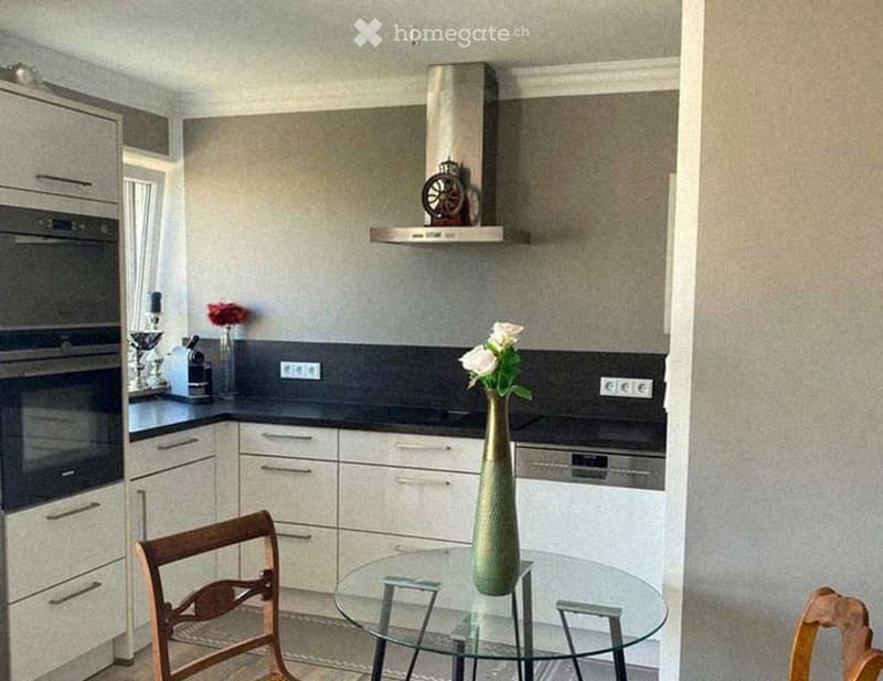 Modern kitchen with white cabinets, stainless steel appliances, a glass dining table with two wooden chairs, and decorative vases on the counter.
