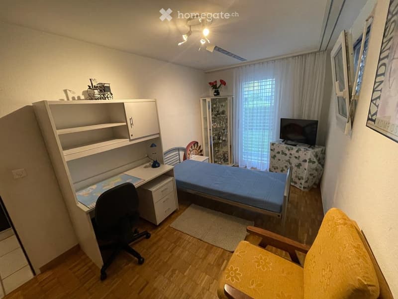 Cozy bedroom with a single bed, desk, office chair, cabinet, small table with TV, and a yellow armchair. The room features wooden flooring, white walls, and a window with curtains.
