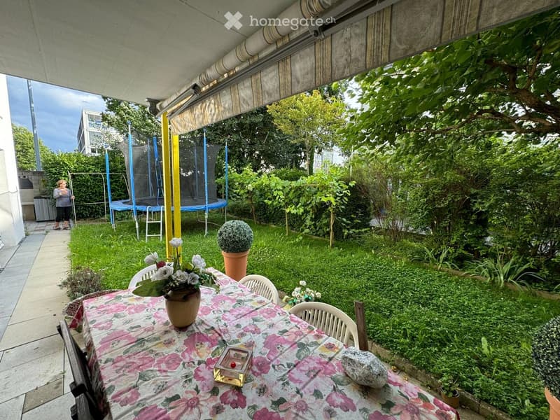Garden area with a table, chairs, and a vase of flowers, surrounded by greenery and a trampoline.
