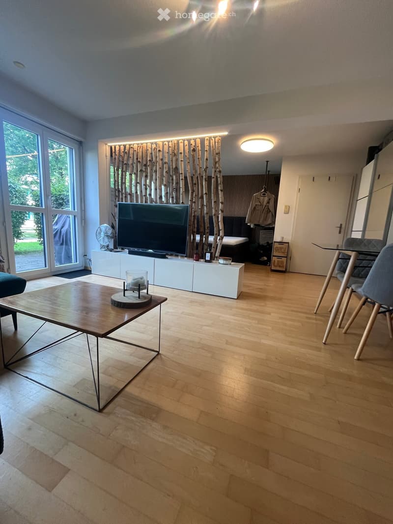 Modern living room with wooden flooring, large windows, a sofa, coffee table, TV, and a fan. The room features a contemporary design with a mix of wooden and metal furniture.