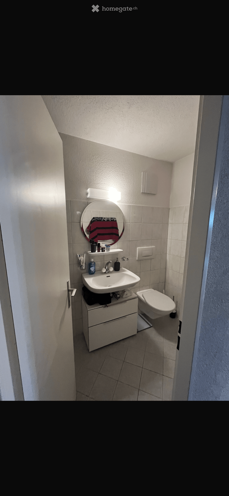 Modern bathroom with a white sink, toilet, round mirror, and storage cabinet. The walls are tiled, and the floor is tiled as well.