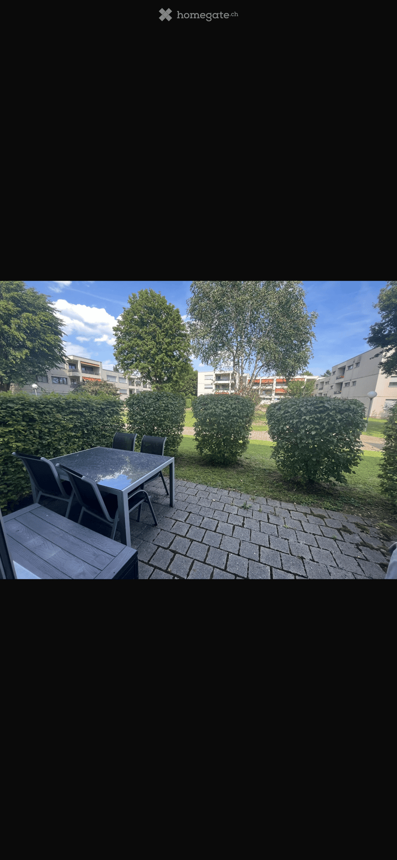 Outdoor patio area with a table and chairs, surrounded by greenery and apartment buildings in the background.