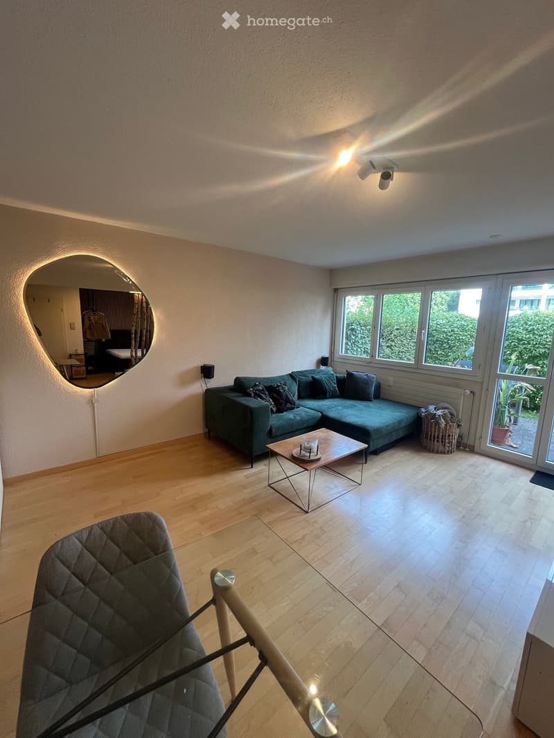 Modern living room with wooden flooring, large windows, a green sectional sofa, a glass coffee table, and a grey quilted chair.