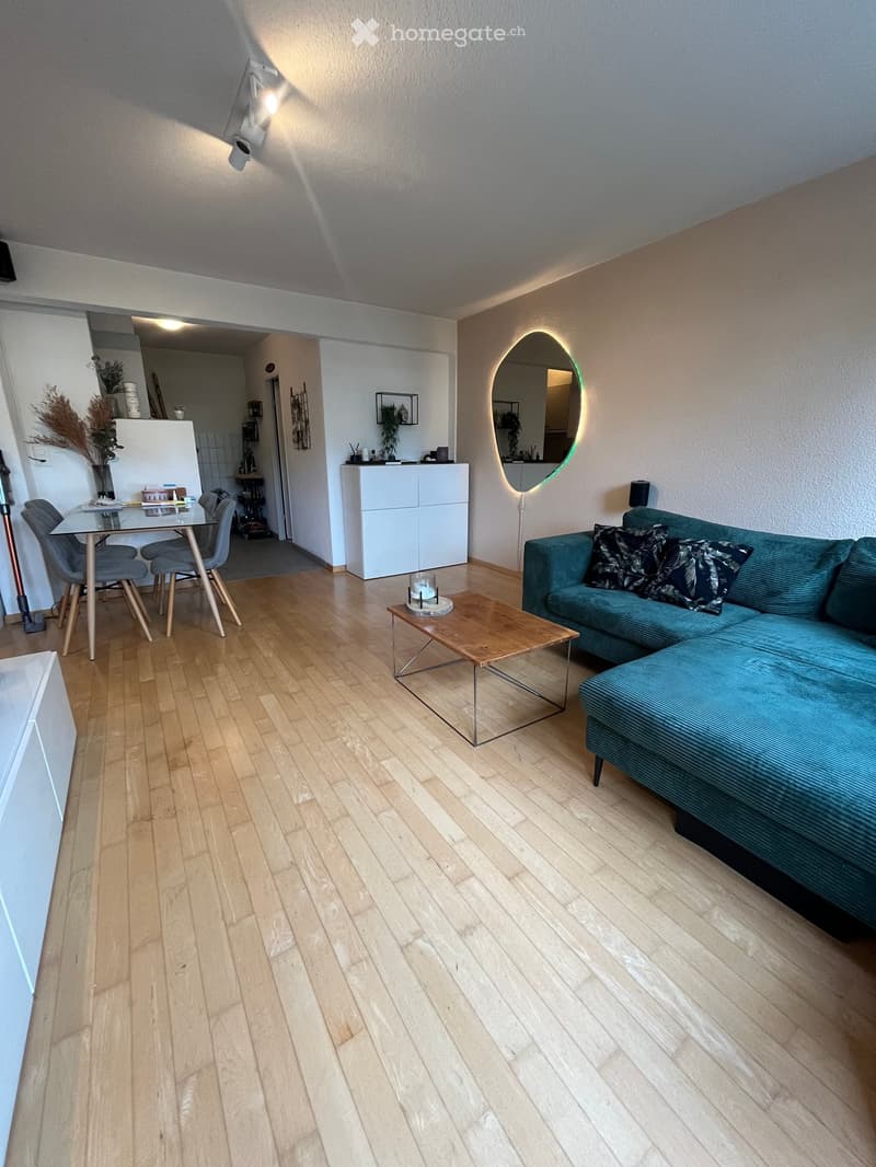 Modern living room with light wooden flooring, a teal sofa, a wooden coffee table, a dining area with a glass table and chairs, and a white cabinet.