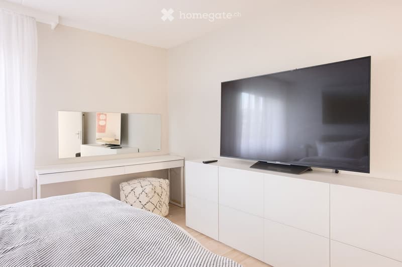 Modern bedroom with a large flat-screen TV on a white cabinet, a neatly made bed with striped bedding, a white dresser with a mirror, and a patterned stool.